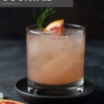 Single low ball glass on a dark gray background filled with ice, grapefruit juice and gin. it is garnished with a fresh wedge of grapefruit and fresh rosemary.