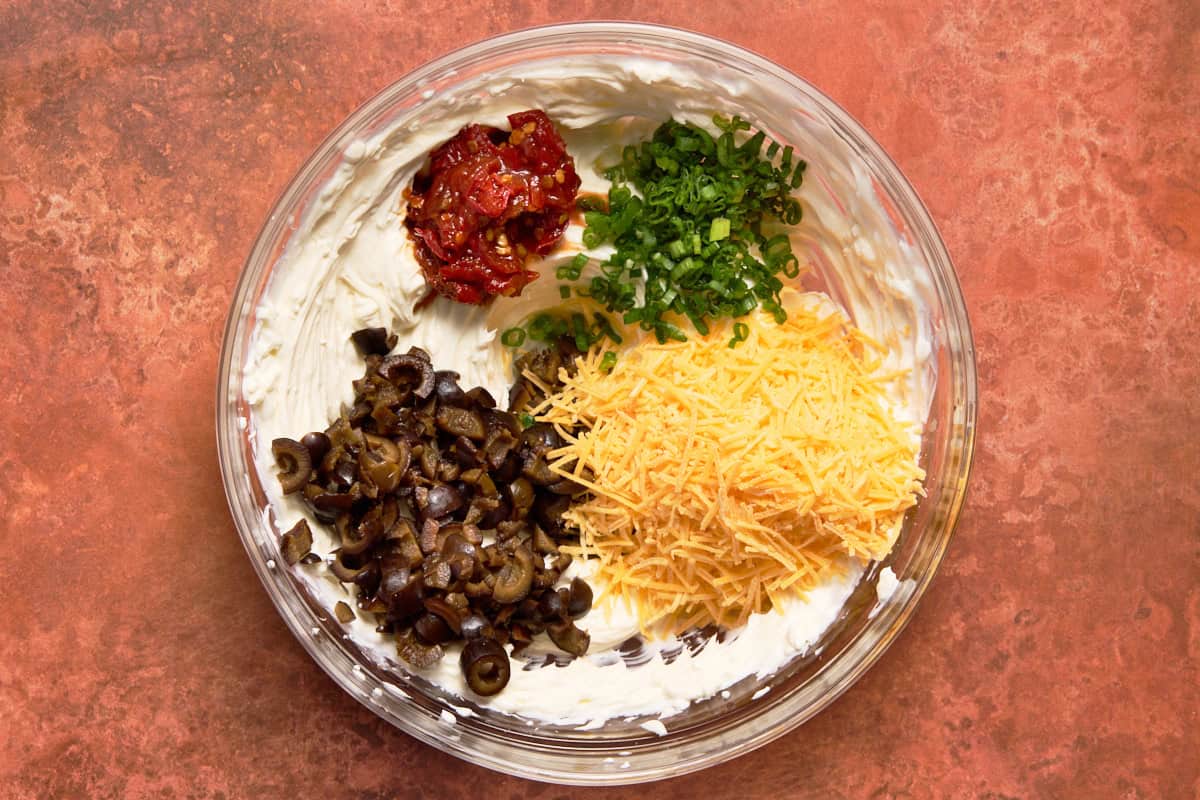 Bowl filled with cream cheese, shredded cheese, olives, green onions and chipotle peppers.